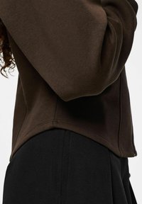 Sweat-shirt en coton marron avec une coupe ample, doté d'un ourlet arrondi et de manches à poignets. Associé à un bas texturé noir.