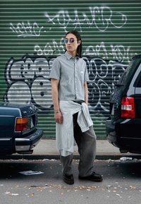 Chemise à manches courtes rayée, pantalon gris oversized et chaussures noires. Accessoires incluent des lunettes de soleil et une longue chemise nouée à la taille.