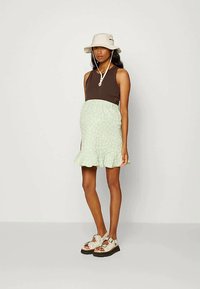 Vestido floral em verde claro com bainha em babados, combinado com um top castanho sem mangas. Acessórios incluem um chapéu de balde bege e sandálias.