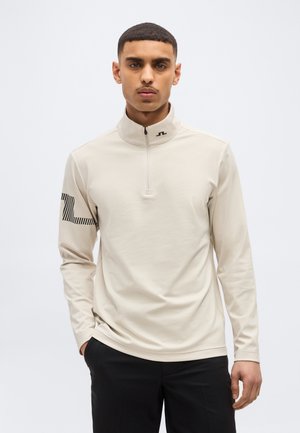 Noor mees, kes kannab beeži värvi kvartszipiga pusa, millel on must logo varrukal ja krael, seisab ühe käega taskus.