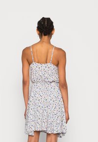 Nainen, jolla on punottu tukka ja joka pukeutunut hihattomaan, ohuilla olkaimilla varustettuun kukkakuosiseen mekkoon, kuvattuna selin yksinkertaisella taustalla.