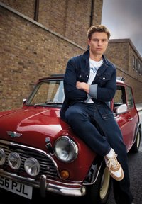 Coche vintage rojo con acentos de cromo; una persona con una chaqueta de mezclilla oscura y pantalones se sienta en el capó, usando zapatillas blancas y marrones.