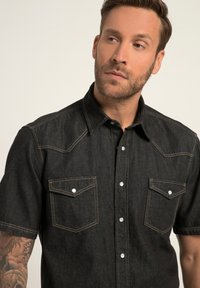 Tumman denim-kankaan lyhythihainen paita nappikauluksella, kahdella rintataskulla, valkoisilla ompeluvaroilla ja terävillä olkapääyksityiskohdilla.