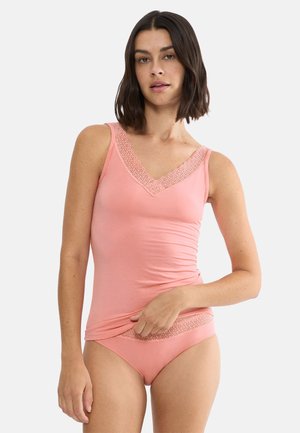 Frau mit schulterlangem dunklem Haar, die ein rosa ärmelloses Oberteil und passende Unterwäsche mit Spitzenbesatz trägt, vor einem einfarbigen Hintergrund stehend.