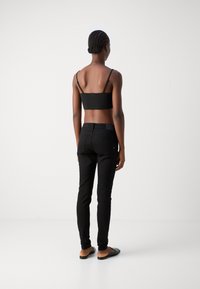 Mujer con el cabello trenzado, vistiendo un top corto negro, jeans negros ajustados y zapatos negros sin cordones, de pie de espaldas contra un fondo blanco liso.