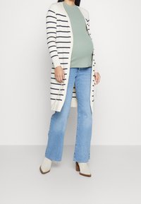 Lång cardigan med öppen framsida i krämfärg med marinblå ränder; parat med en grön topp och blå utsvängda jeans; har klackade ankelskor.