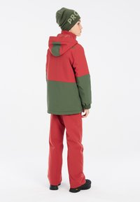 Rote und grüne Jacke mit Kapuze und elastischen Bündchen, kombiniert mit roten Hosen. Eine grüne Mütze hat kontrastierende Schriftzüge.