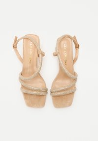 Sandales à talons hauts beiges avec une texture en daim, présentant trois brides métalliques et un design à bout carré. Logo doré sur la semelle intérieure.