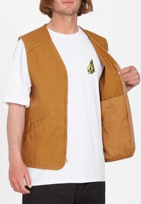 Homme portant un t-shirt blanc avec un petit motif géométrique jaune, montrant un gilet marron sans manches ouvert avec une doublure intérieure et des poches.