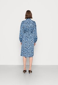 Vestido de estampa de onça azul com mangas longas, cintura ajustada e comprimento até o joelho. Tecido liso com um leve brilho. Usado com mules pretos de salto.