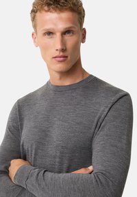 Langarmiges graues Strickshirt mit rundem Ausschnitt, glatter Textur und engen Ärmeln, getragen von einem Modell mit verschränkten Armen.