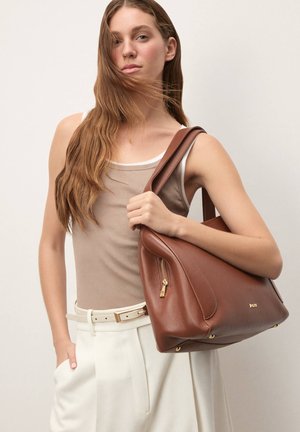 Brown leather handbag with a structured shape, featuring a side zip pocket and gold-tone accents. Model holding bag, wearing a beige top and white pants.