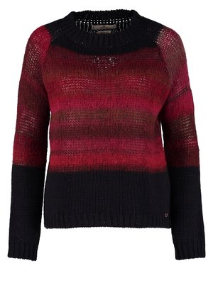 Personne portant un pull dégradé rouge et noir, un jean skinny bleu délavé et des bottines noires brillantes, debout avec une main dans la poche.
