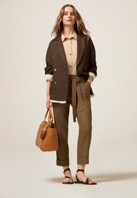 Bruine dubbelgebreide blazer, beige shirt, olijfgroene high-waisted broek met ceintuur, bruine handtas en zwarte sandalen met een gouden accent.