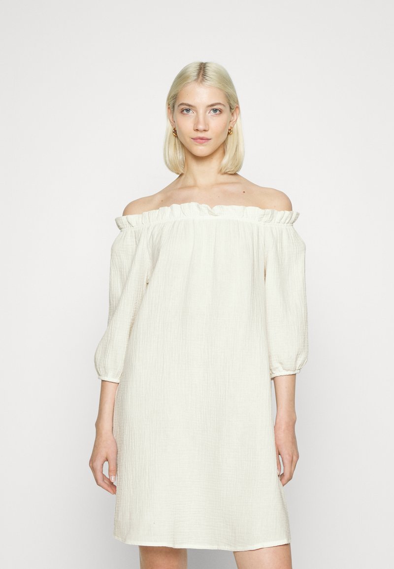 Vestido creme com ombros à mostra, decote com babados, mangas de três quartos e tecido texturizado, apresentando um corte solto e comprimento até o joelho.