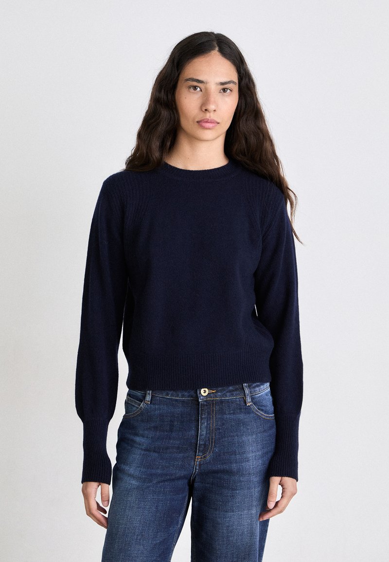 Marineblauer Strickpullover mit Rundhalsausschnitt, gerippten Bündchen und Saum. Getragen mit blauen Jeans, die eine enganliegende Passform und glatte Textur aufweisen.