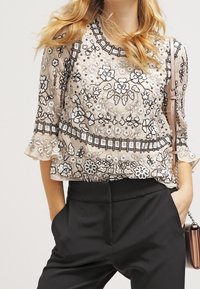 Femme portant un chemisier beige transparent avec une broderie florale noire et blanche, un pantalon noir taille haute, les mains dans les poches, tenant un petit sac à main.