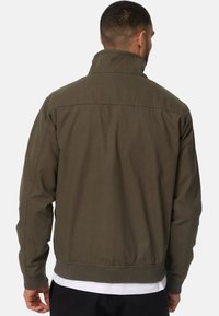 Veste bomber vert olive avec col montant, poignets et taille côtelés. Fabriquée dans un tissu lisse, elle présente une coupe décontractée et un design minimaliste.