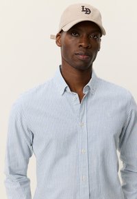 Μπεζ καπέλο του μπέιζμπολ με κεντημένα γράμματα "LD", φορεμένο πάνω από πουκάμισο με κουμπιά σε ανοιχτό μπλε και λευκές ρίγες. Διακρίνονται καθαρά η υφή του υφάσματος και οι λεπτομέρειες των κουμπιών.