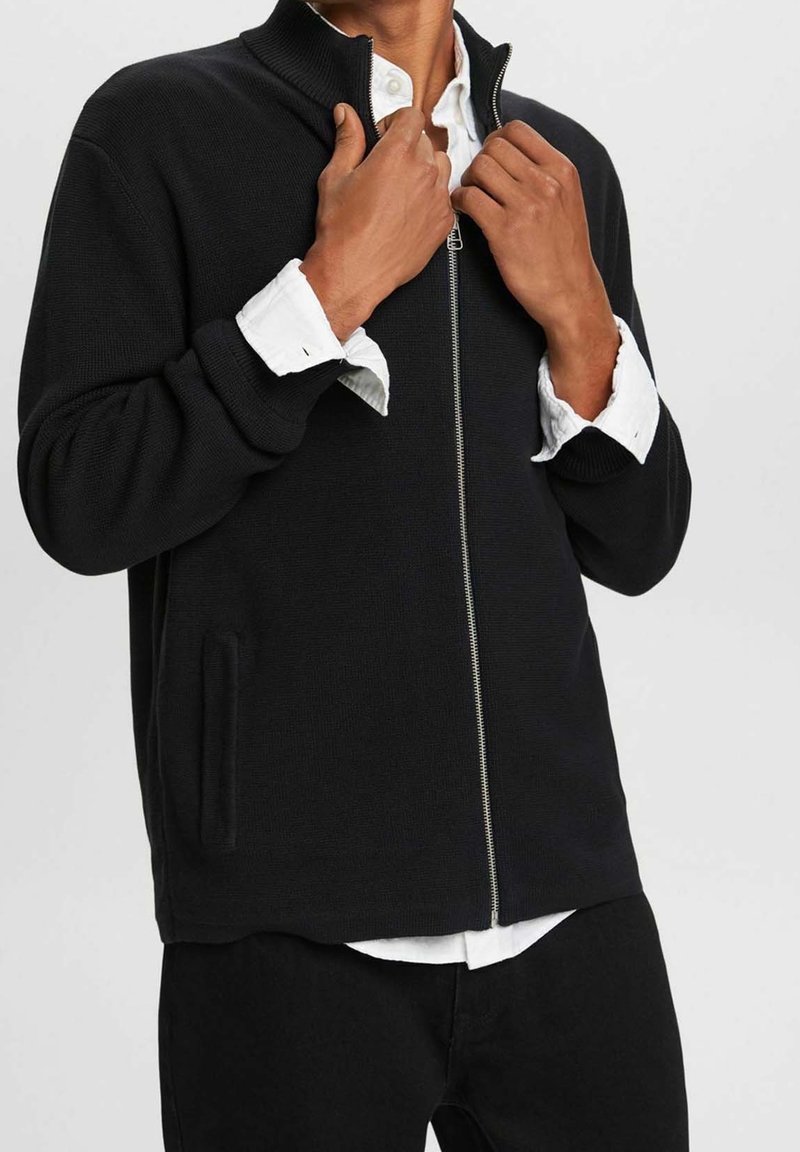 Homme ajustant le col d'un pull noir zippé porté par-dessus une chemise blanche boutonnée et un pantalon noir sur fond uni.