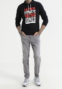 Felpa nera con cappuccio con testo audace in rosso e bianco, abbinata a jeans grigio chiaro e scarpe da ginnastica bianche con dettagli neri.