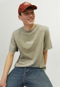 Kortærmet, rundhalset grøn t-shirt med en falmet finish og hvid "Originals Studio" tryk, parret med blå denim jeans og en rustfarvet kasket.