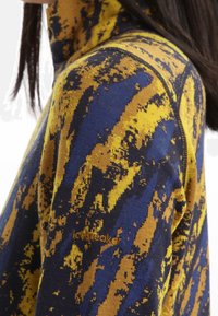 Fleece-Pullover in blau-gelbem abstraktem Muster, mit hohem Kragen, Reißverschluss und Logodetail in goldfarbener Stickerei.