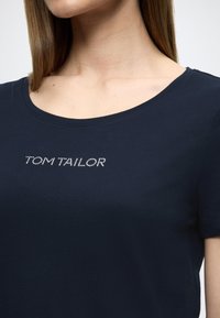 Μπλουζάκι από βαμβάκι σε ναυτικό μπλε χρώμα με στρογγυλή λαιμόκοψη και κοντά μανίκια. Διακοσμημένο με την επιγραφή «TOM TAILOR» από κρυσταλλικές διακοσμήσεις στο στήθος.