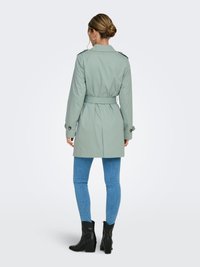 Trench-coat vert clair avec une taille ceinturée, des épaulettes et des manches longues, assorti à un jean skinny bleu et des bottes à talons noires.