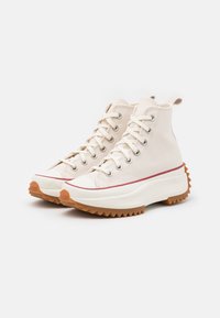 Płócienne sneakersy typu high-top w kolorze kremowym z teksturowaną gumową podeszwą, czerwoną akcentującą paskiem i tradycyjnym wzorem sznurowania.