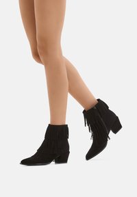 Schwarze Wildleder-Ankle Boots mit Fransenverzierung, spitzem Zehenbereich und niedrigem Blockabsatz. Reißverschluss an der Seite für ein einfaches Anziehen.