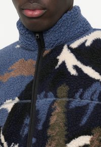 Polarowa kurtka z wysokim kołnierzem w niebieskie, brązowe i kremowe abstrakcyjne wzory. Posiada czarny zamek błyskawiczny oraz kontrastowe detale szwów.