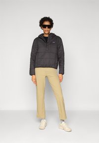 Zwarte pufferjas met een capuchon, voorzien van een gewatteerde textuur en een drukknoopsluiting aan de voorkant, gecombineerd met beige wijde broek en lichte sneakers.