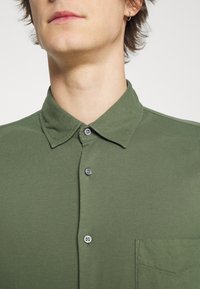 Oliverově zelená košile s límečkem a pěti knoflíky, texturovaná tkanina a náprsní kapsa na levé straně.
