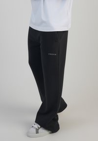 Melni sporta bikses ar brīvu piegriezumu, ar smalku SIKSILK logo kreisajā pusē, apvienotas ar baltām sporta kurpēm.