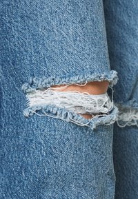 Denim azul desgastado con bordes deshilachados y un agujero recortado que revela hilos blancos. La textura es suave con un acabado de lavado medio.