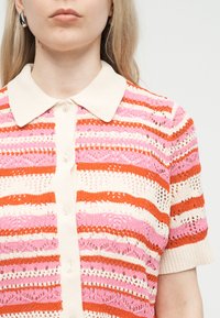 Femme portant un polo en maille à rayures avec des motifs horizontaux roses, rouges et blancs et des manches courtes, sur un fond clair uni.