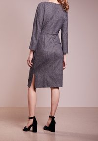 Robe grise à motifs, avec manches longues, fente sur le côté, taille cintrée et tissu lisse. Associée à des chaussures à talons bloc noires.