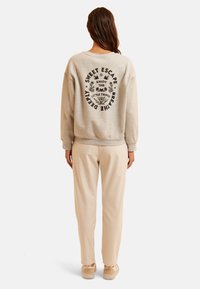 Sudadera de algodón gris claro con diseño de texto negro en la parte posterior, combinada con pantalones beige y zapatos claros. Ajuste relajado, estilo simple.