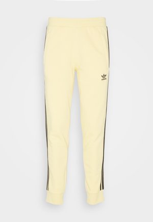 Jasnożółte spodnie dresowe Adidas z czarnymi trzema paskami po bokach i logo na lewej udzie, elastycznym pasem i ściągaczami przy kostkach.