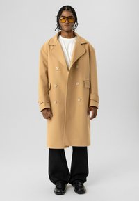 Beige dubbelgeknoopte jas met grote kraag en twee zakken. Gedragen over een wit overhemd, gecombineerd met een zwarte broek en zwarte schoenen.