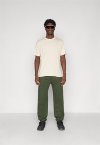 Beige kortermet t-skjorte, grønne joggebukser og svarte sneakers. Modellen har på seg mørke solbriller. Enkelt design, løs passform, ingen mønstre.
