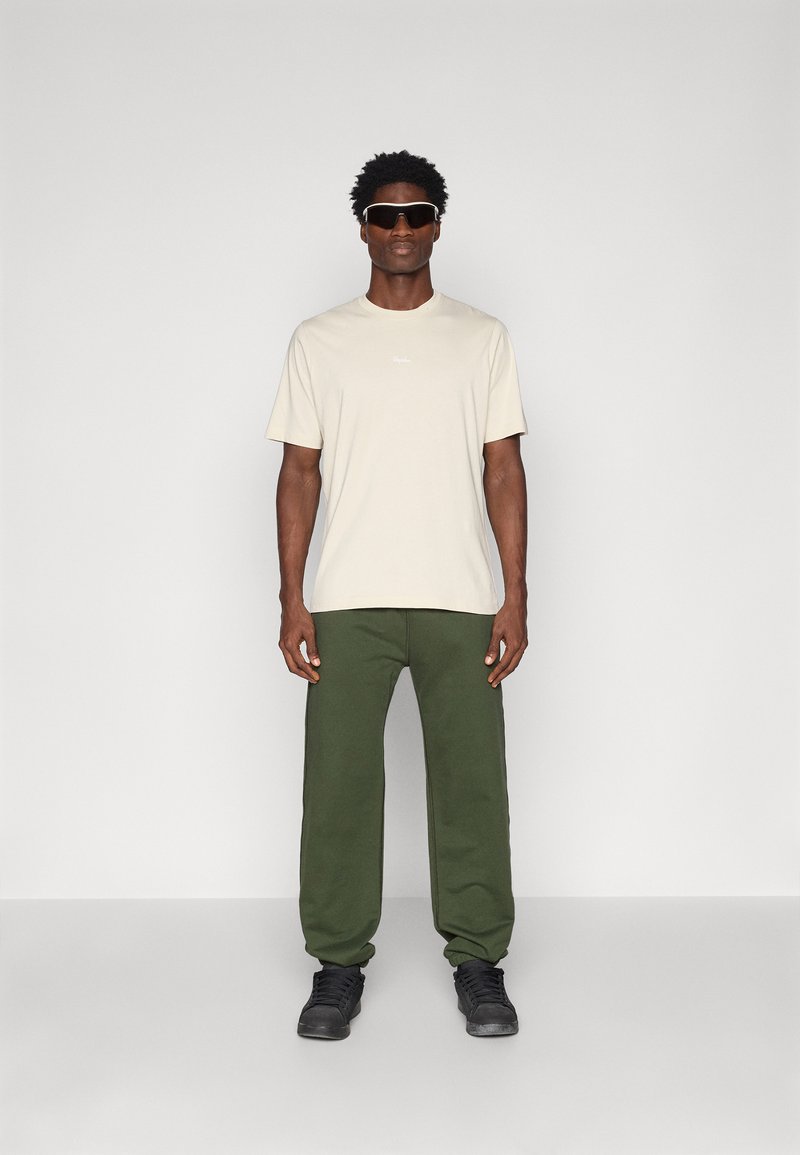 Beige T-Shirt mit kurzen Ärmeln, grüne Jogginghose und schwarze Turnschuhe. Das Modell trägt eine dunkle Sonnenbrille. Einfaches Design, entspannte Passform, keine Muster.