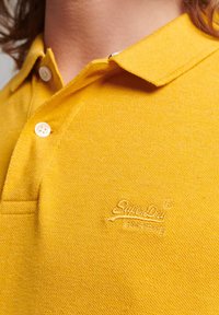 Gul polo shirt lavet af tekstureret stof, med en tre-knap lukning og broderet logo på brystet. Korte ærmer.