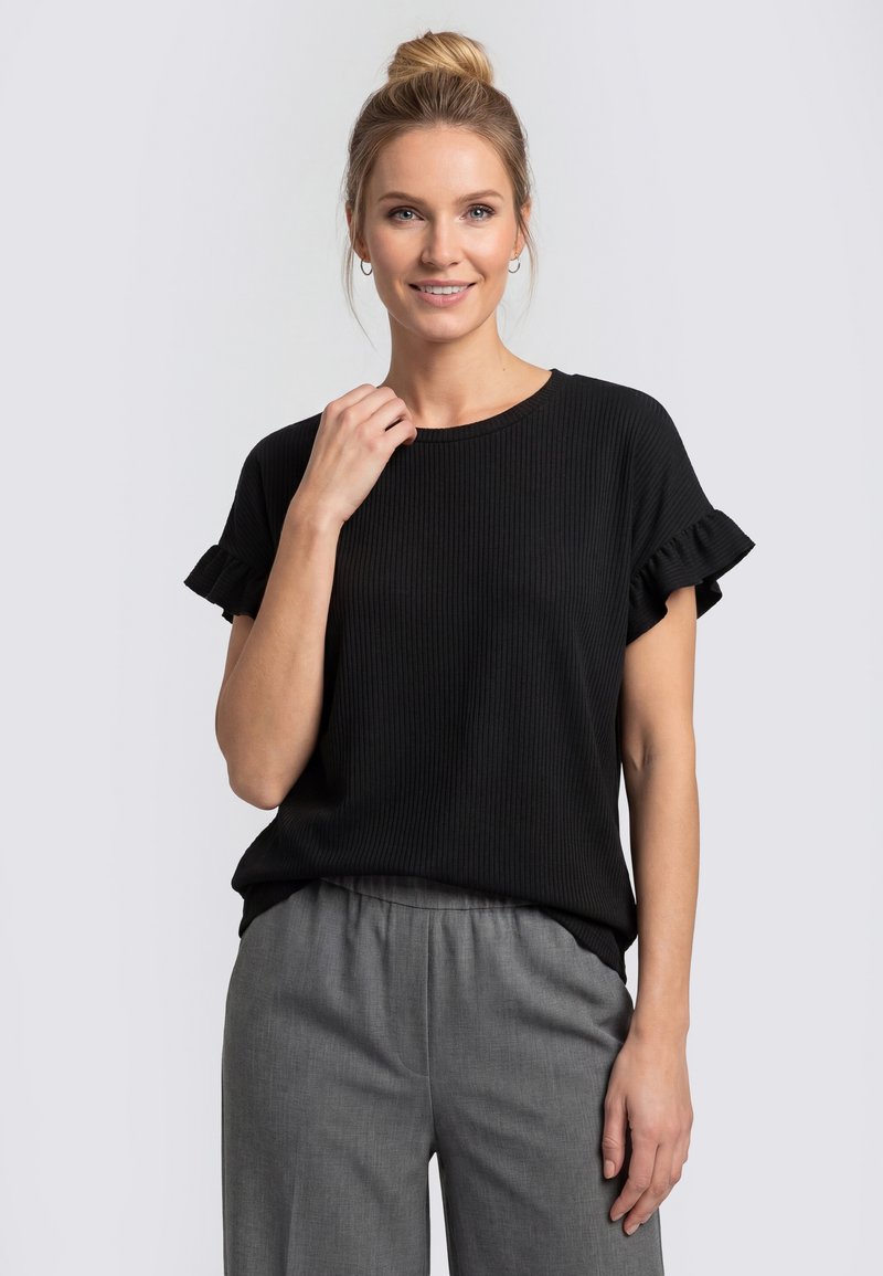Femme aux cheveux blonds en chignon, portant un haut noir à manches courtes avec des manches à volants et un pantalon gris, souriante et touchant son cou.