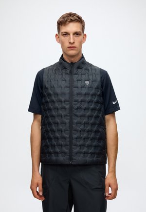 Jeune homme aux cheveux courts portant un gilet matelassé noir zippé par-dessus un t-shirt noir Nike à manches courtes et un pantalon noir sur fond blanc.