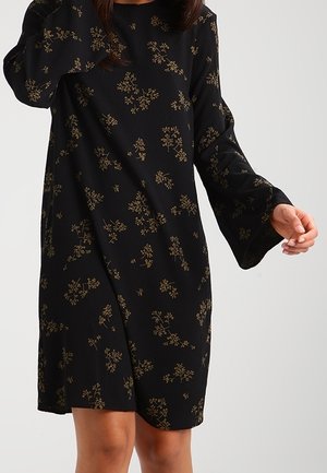 Vrouw met een zwarte jurk met lange mouwen en kleine beige bloemenpatronen, staand tegen een effen lichte achtergrond.