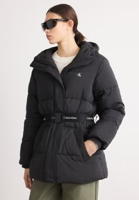 Crna skijaška jakna s kapuljačom, koja ima remen oko struka i Calvin Klein logo. Dizajn ima prošivenu teksturu i bočne džepove.