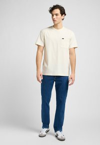 Beżowa koszulka z krótkim rękawem z kieszonką na piersi, połączona z niebieskimi dżinsami i białymi trampkami w czarne i szare paski.