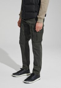 Zelene cargo hlače s više džepova i patent zatvarača, uparene s crnim tenisicama. Outfit sadrži podstavljeni crni prsluk i laganu košulju.